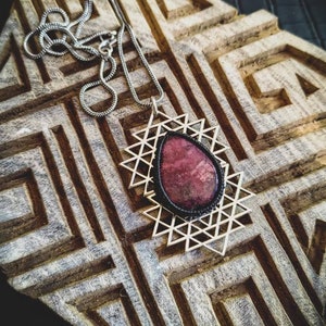 Peut inclure: Un collier en chaîne argent avec un pendentif géométrique orné d'une pierre précieuse rose. Le pendentif est entouré d'un cordon noir et d'un cadre en fil d'argent.