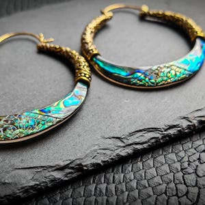 Könnte beinhalten: Ein Paar goldfarbene Creolen mit schillernden Abalone-Muscheleinlagen. Die Ohrringe haben ein detailliertes, strukturiertes Design auf dem goldenen Teil und eine geschwungene, farbenfrohe Muscheleinlage. Die Ohrringe werden auf einer dunklen, strukturierten Oberfläche präsentiert.