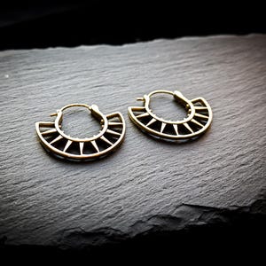 May include: A pair of gold hoop earrings with a geometric design. The earrings are made of metal and have a circular shape with a pointed edge.