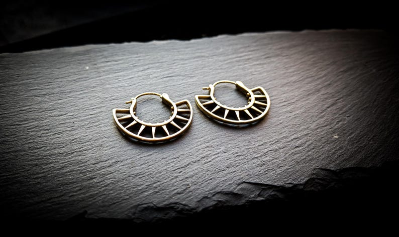 May include: A pair of gold hoop earrings with a geometric design. The earrings are made of metal and have a circular shape with a pointed edge.