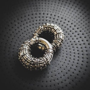 May include: Two silver rings with intricate designs and a textured surface. The rings are linked together and are on a black background.