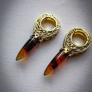 May include: A pair of gold-toned earrings with amber-colored teardrop-shaped stones. The earrings have a detailed, ornate design.