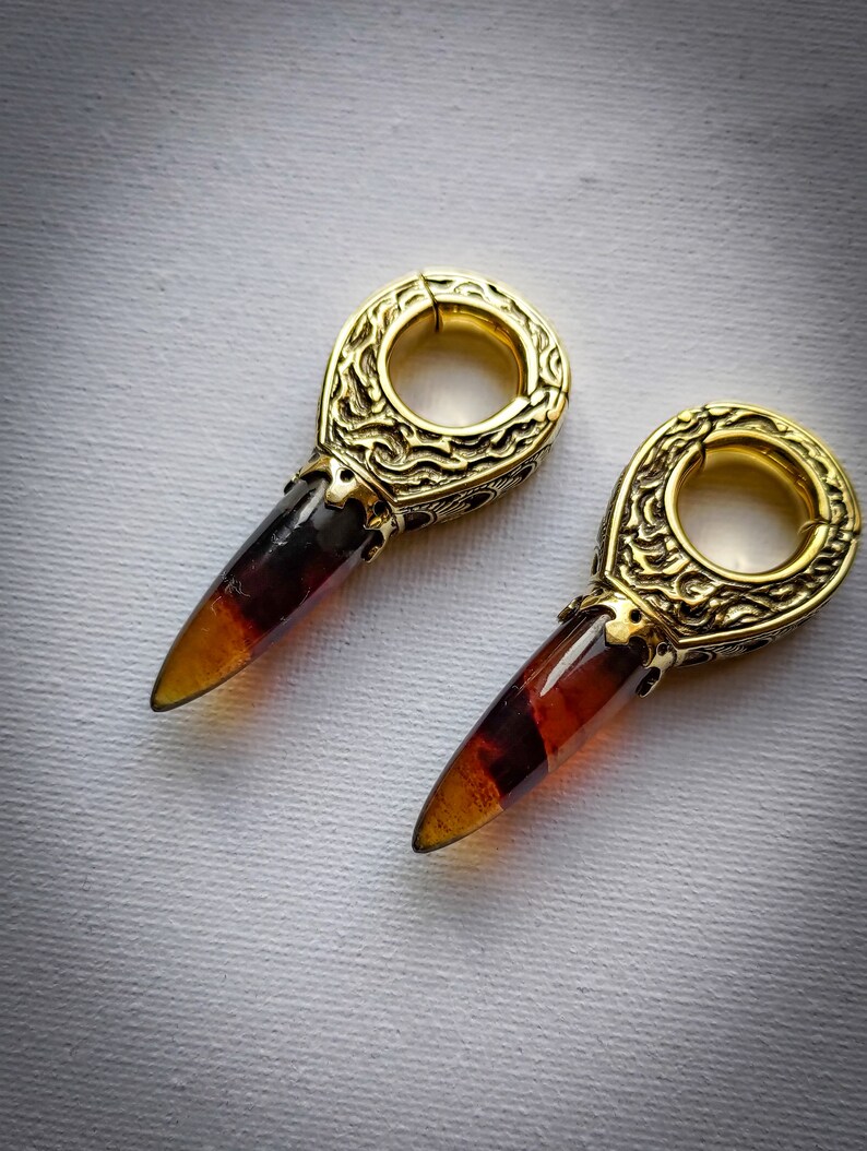May include: A pair of gold-toned earrings with amber-colored teardrop-shaped stones. The earrings have a detailed, ornate design.