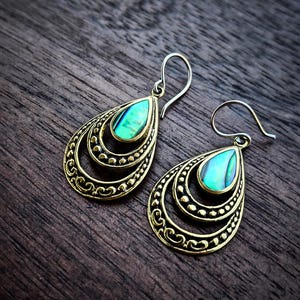 Peut inclure: Une paire de boucles d'oreilles pendantes en forme de goutte, ornées d'incrustations de nacre d'ormeau aux reflets verts et bleus, serties dans des cadres en métal doré vieilli. Elles sont dotées de crochets en métal argenté.