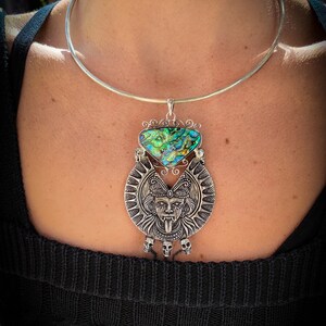 May include: A silver choker necklace with a large pendant featuring a silver skull with a tongue sticking out and a green and blue abalone shell.