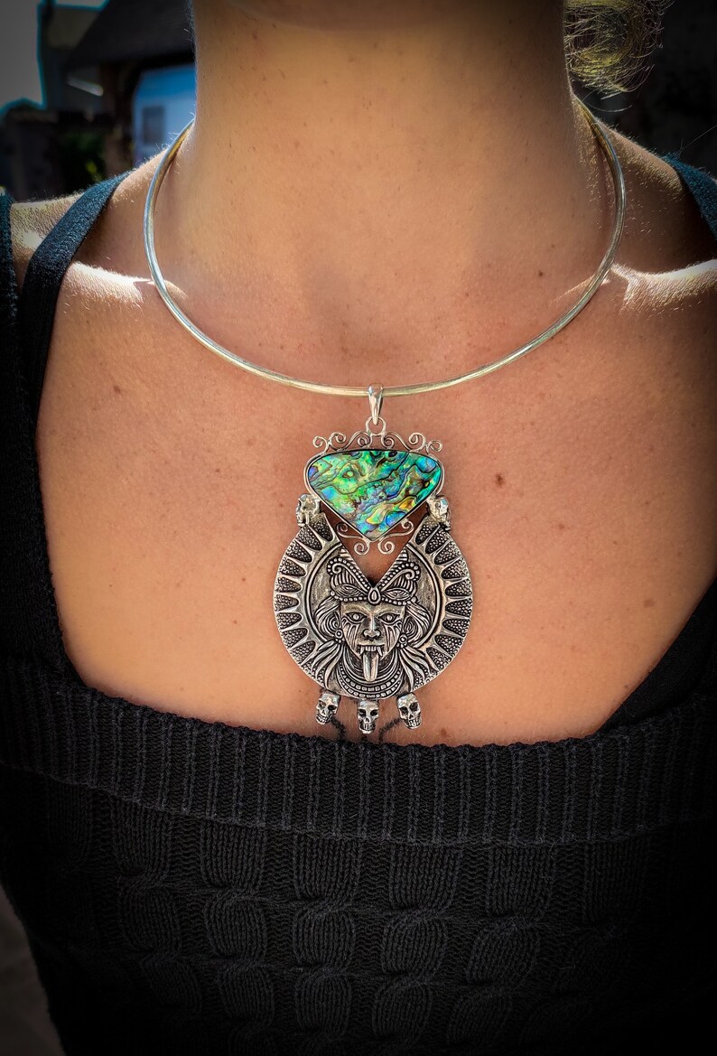 May include: A silver choker necklace with a large pendant featuring a silver skull with a tongue sticking out and a green and blue abalone shell.