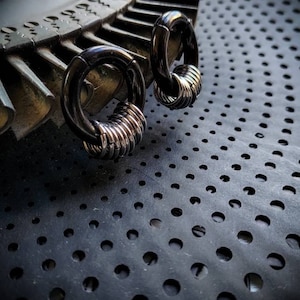 May include: Two black hoop earrings with silver rings stacked on top. The earrings are on a black surface with a pattern of small holes.