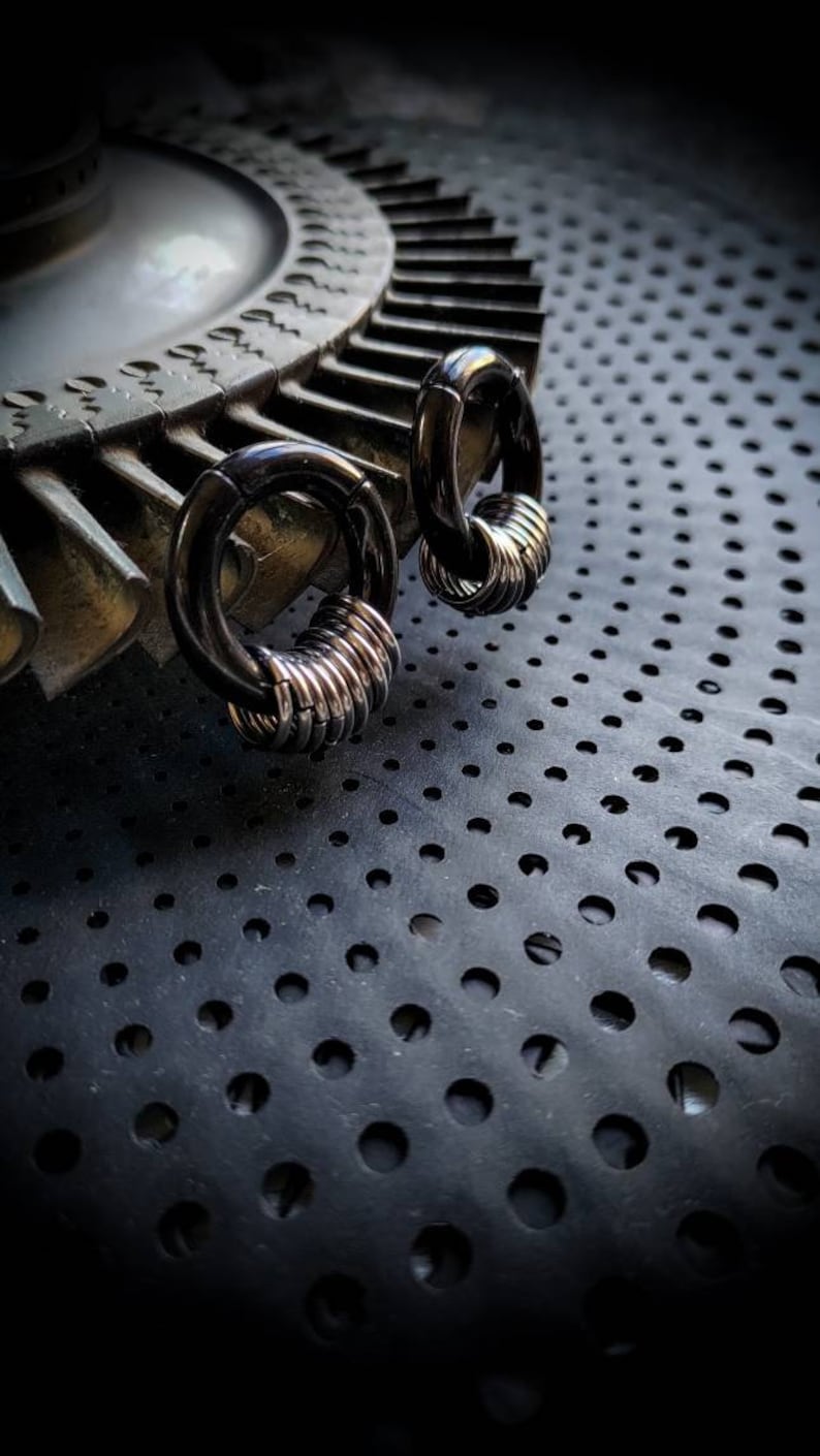 May include: Two black hoop earrings with silver rings stacked on top. The earrings are on a black surface with a pattern of small holes.