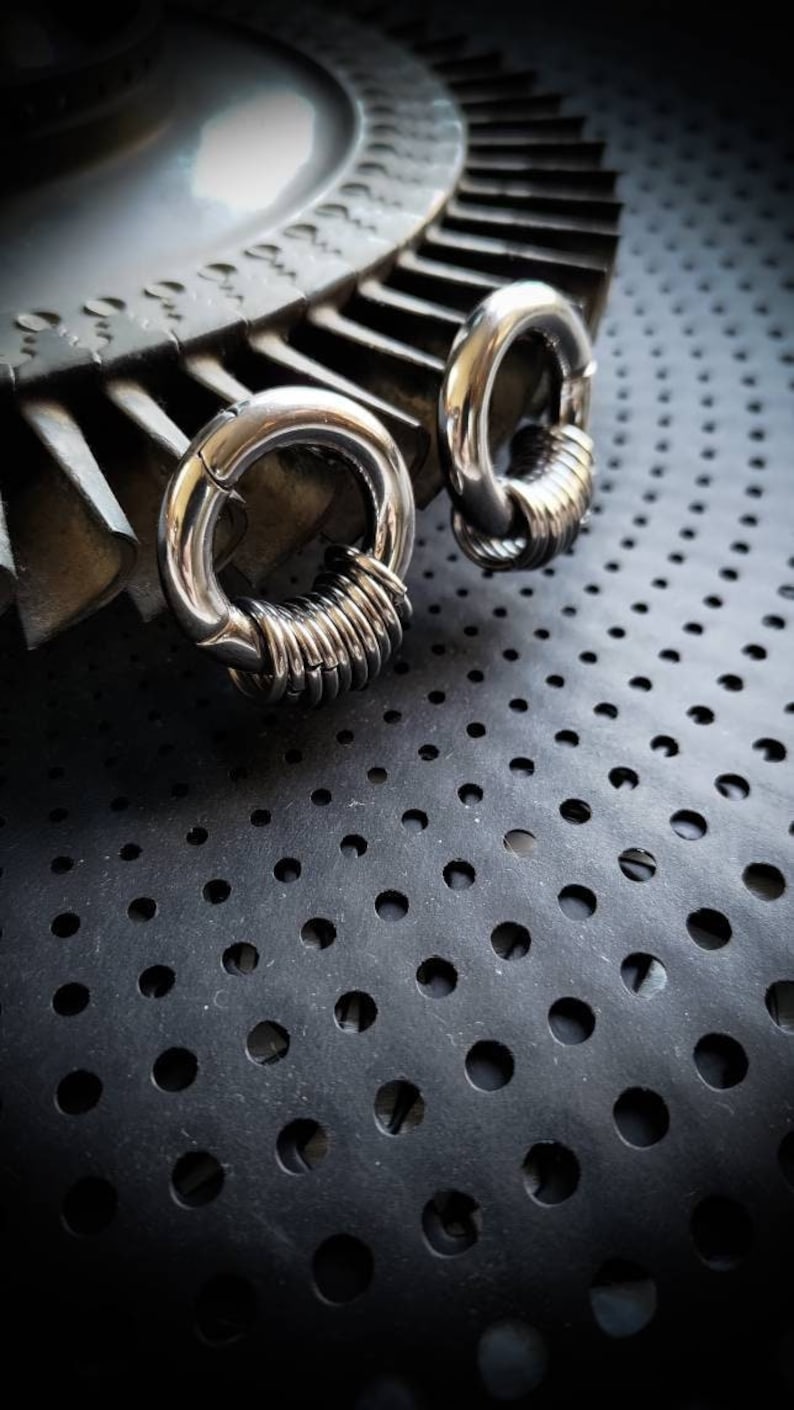 May include: Two silver hoop earrings with a coiled design. The earrings are on a black surface with a pattern of small holes.