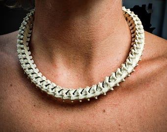 May include: Off-white necklace crafted to resemble a spine, featuring individual vertebrae with black accents. The necklace is worn around the neck, showcasing a unique and edgy design. The necklace is a statement piece.