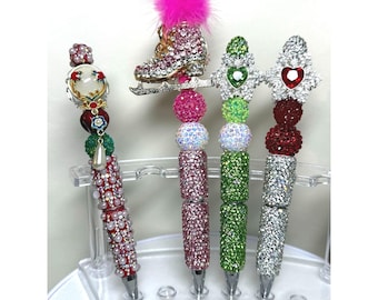 May include: Four decorative pens with jewel-encrusted designs. One pen features a pink ice skate with a fluffy pink pom-pom. Other pens have jewel-toned accents, including hearts and a pearl-like design. The pens are displayed on a clear plastic stand.