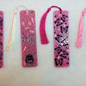 May include: Four glittery pink resin bookmarks with tassels. One has elephant graphics and the text "MOM LIFE CHAOS DOMINATES". Another says "Nurse Life" with a leopard print design. The others have butterfly and heart designs.
