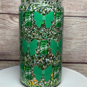 May include: A clear, glitter-filled tumbler with a screw-on lid. The tumbler is decorated with green bows and Sprite cans. The glitter is a mix of green, white, and other colors. The tumbler is cylindrical in shape.
