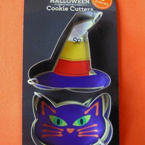 May include: A two-piece set of Halloween cookie cutters on a black display card. One cutter is shaped like a witch hat with yellow, orange, and purple sections. The other is shaped like a purple cat face with green eyes and orange whiskers.