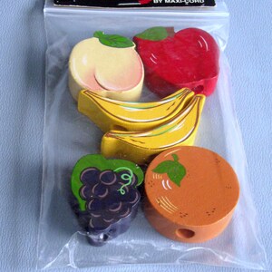 May include: A clear plastic bag containing six wooden macrame beads shaped like fruit, including a peach, apple, two bananas, grapes, and an orange. The header card reads Maxi-Shapes A Macrame Bead BY MAXI-CORD. Each bead features a central hole.