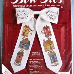 May include: A cross-stitch kit for a door decoration. The kit features a white bow with the text "Bow Ties" and "There's no place like home." The design includes cross-stitched houses in various colors. Finished size: 11" x 17".