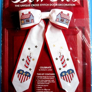 May include: A cross-stitch door decoration kit featuring a patriotic bow tie design. The bow tie has a white base with red trim and includes cross-stitched houses, fireworks, stars, and a heart with stars and stripes. The kit includes instructions.