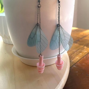 May include: A pair of dangle earrings with blue butterfly wings and pink earplugs hanging from a black chain.