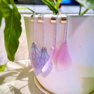 May include: Four pairs of dangle earrings with delicate silver chains and translucent wings. Two pairs are light blue and two pairs are light pink.