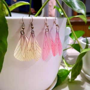 May include: Two pairs of earrings with delicate butterfly wings. One pair is a pale yellow with brown veins, the other is a soft pink with white veins. The earrings are hanging from a white ceramic pot.