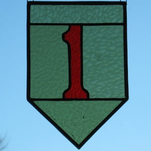May include: A stained glass window hanging with a chain. The window is shaped like a shield and is green with a red number one in the center.