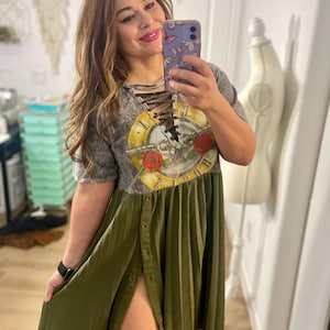 May include: A grey and olive green dress with a graphic print. The top has a lace-up neckline and a clock design with roses. The skirt is olive green, pleated, and button-down. The dress is worn with a black watch.