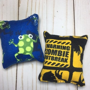 May include: Two small fabric bean bags. One is blue with a green frog design and the other is yellow with black zombie warning signs and silhouettes.