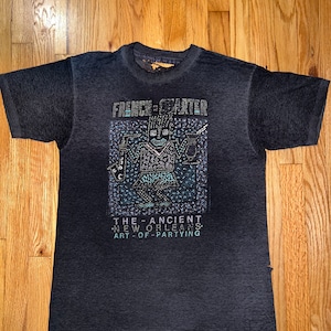 May include: A dark gray t-shirt with a graphic print of a stylized figure and the text "French Quarter The Ancient New Orleans Art of Partying".