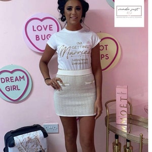 May include: A white t-shirt with gold glitter text that reads "I'm Getting Married" with a heart and arrow and the text "Lucys hende Liverpool 2121". The person is wearing a white skirt with gold trim and a gold belt.