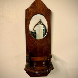 May include: A vintage wooden wall-mounted mirror with an oval mirror and a small shelf. The frame is a dark brown color with a decorative top and a shelf with carved details. The mirror reflects a room.
