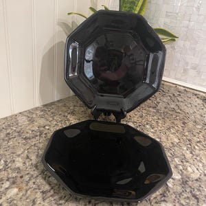 May include: Two black octagonal serving dishes. The top dish is elevated on a black stand, while the bottom dish rests on a speckled countertop. The dishes have a glossy finish and reflect light.