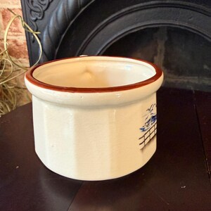 May include: Cream-colored ceramic container with a brown rim. The cylindrical pot has a blue and white illustration on the side. The container is sitting on a dark brown surface.