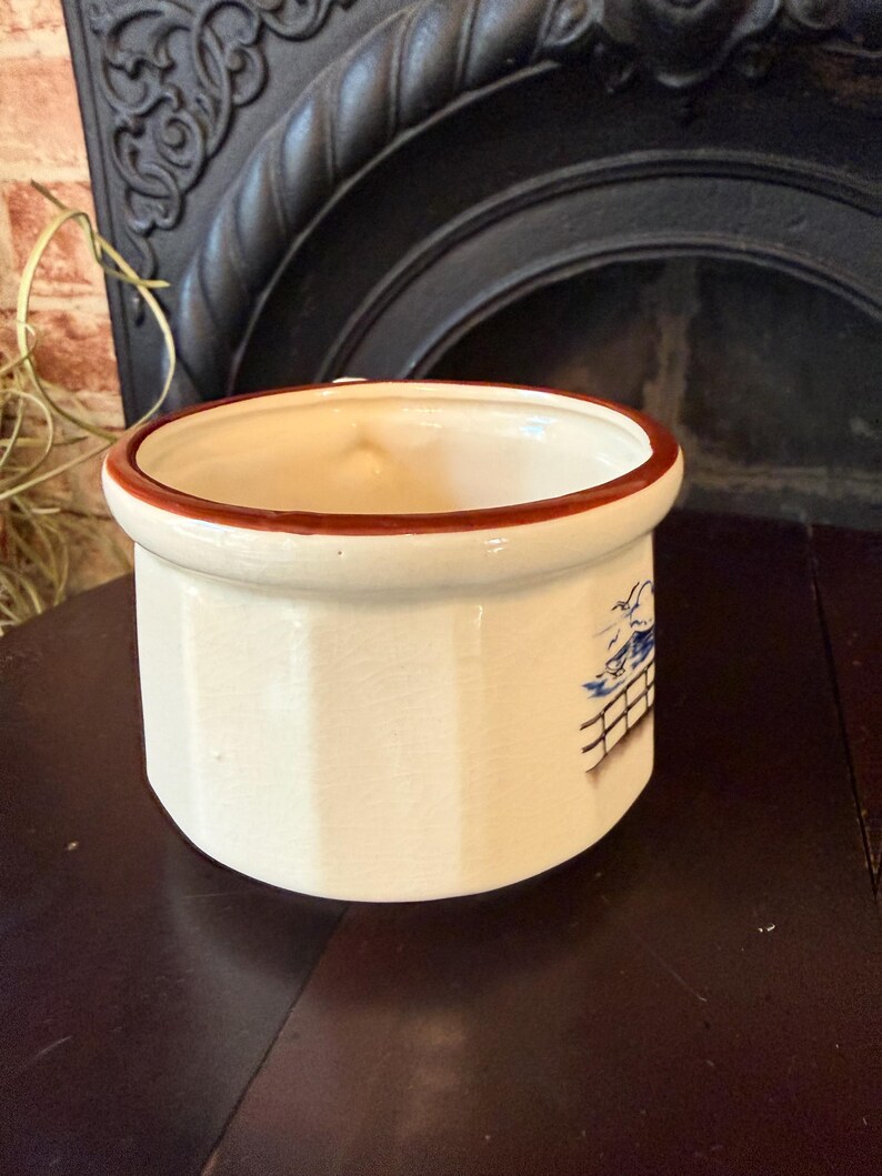 May include: Cream-colored ceramic container with a brown rim. The cylindrical pot has a blue and white illustration on the side. The container is sitting on a dark brown surface.
