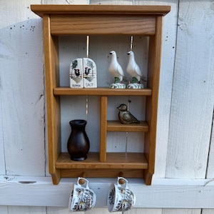 May include: A wooden wall shelf with multiple compartments, holding decorative items. The shelf displays a salt and pepper shaker, two white dove figurines, a small bronze bird, a dark vase, and two decorative mugs. The shelf is made of light brown wood.