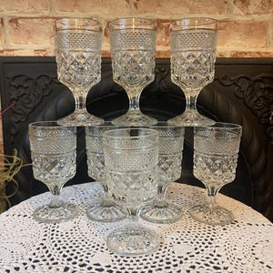 May include: A collection of eight clear, vintage-style water goblets. Each goblet features a diamond-patterned design on the bowl and a stemmed base. The glasses are arranged on a white, lace-like tablecloth, suggesting a classic or antique aesthetic.
