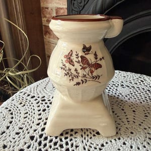 May include: Cream-colored ceramic decorative stove-shaped container with a brown rim and handle. The container features a butterfly and floral design in shades of brown and pink. The base is a rectangular shape with a decorative pattern.