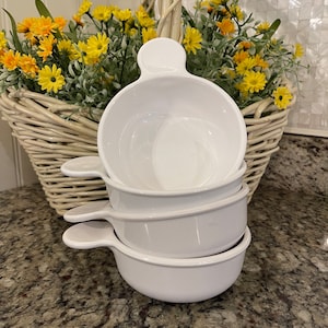May include: A stack of four white soup bowls with handles. The bowls are round and have a simple, clean design. A wicker basket filled with yellow flowers and greenery is in the background.