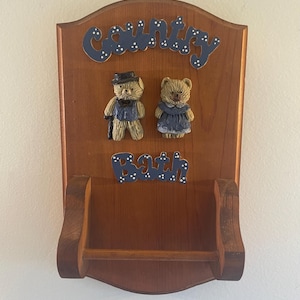 May include: Wooden country-themed wall shelf with the word "Country" in blue script with white polka dots. Two teddy bear figurines are affixed to the wood. The word "Bath" is also in blue script.