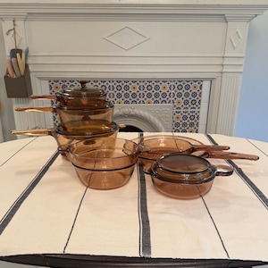 May include: A collection of amber-colored glass cookware including a stack of three saucepans with lids, a wide shallow skillet, and a small round casserole dish with a lid. Each piece features integrated glass handles and a translucent finish.
