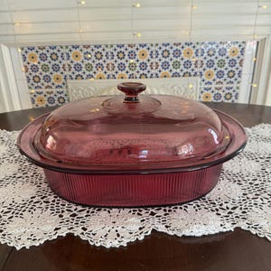 May include: A vintage, oval-shaped, ruby-red glass casserole dish with a matching lid. The dish has a ribbed design and a clear glass lid with a knob handle. The dish is sitting on a white lace doily on a dark wooden table.