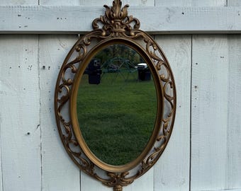 May include: An oval-shaped, antique-style mirror with a gold-colored ornate frame. The mirror is hanging on a white picket fence. The frame features decorative scrollwork and a floral motif at the top and bottom.