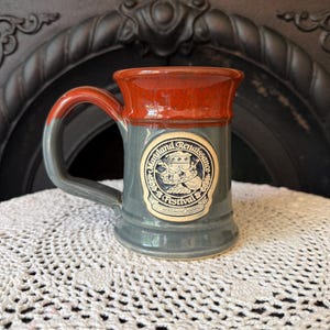 May include: A ceramic mug with a two-tone glaze featuring a rust-orange rim and a grey base. A circular emblem on the front displays the text Maryland Renaissance Festival and Let Merriment Abound, surrounding an illustration of a goblet and crown.