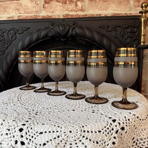 May include: Six vintage wine glasses with frosted gray bowls and gold bands. The glasses have brown stems and sit on a white crocheted tablecloth. The background includes a black fireplace and a brick wall.