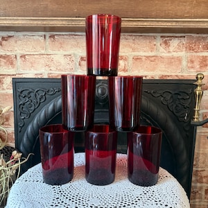 May include: A stack of seven ruby red drinking glasses. The glasses are cylindrical with a slightly flared rim. The glasses are arranged on a white lace tablecloth, with a brick wall and wooden shelf in the background.