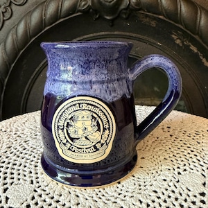May include: A deep blue ceramic mug with a lighter blue glazed rim and a handle. The front features a circular cream-colored emblem with the text Maryland Renaissance Festival and Let Merriment Abound, surrounding an illustration of a goblet.