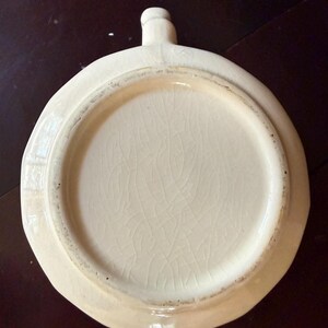 May include: Off-white ceramic dish with a handle. The dish has a circular shape with a slightly irregular edge. The interior has a cracked glaze pattern. The handle is small and rounded.