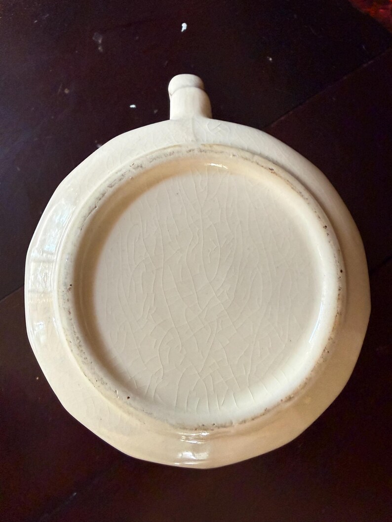 May include: Off-white ceramic dish with a handle. The dish has a circular shape with a slightly irregular edge. The interior has a cracked glaze pattern. The handle is small and rounded.