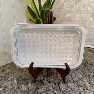 May include: A clear, rectangular glass baking dish with a textured pattern, resting on a dark wooden stand. The dish is empty and sits on a speckled countertop, with a plant in the background.
