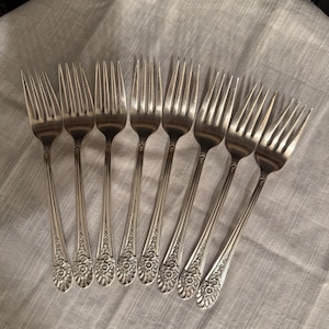 May include: A set of eight silver-toned metal forks arranged in a fan pattern. Each fork features a long, slender handle with a detailed floral and leaf embossed design at the base and four tines at the top, reflecting a polished metallic finish.