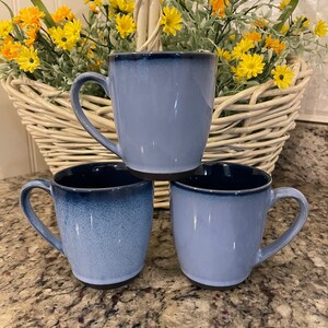 May include: Three blue ceramic mugs with dark blue rims and handles are arranged on a speckled countertop. A white wicker basket filled with yellow flowers and greenery is in the background, creating a spring-like aesthetic.